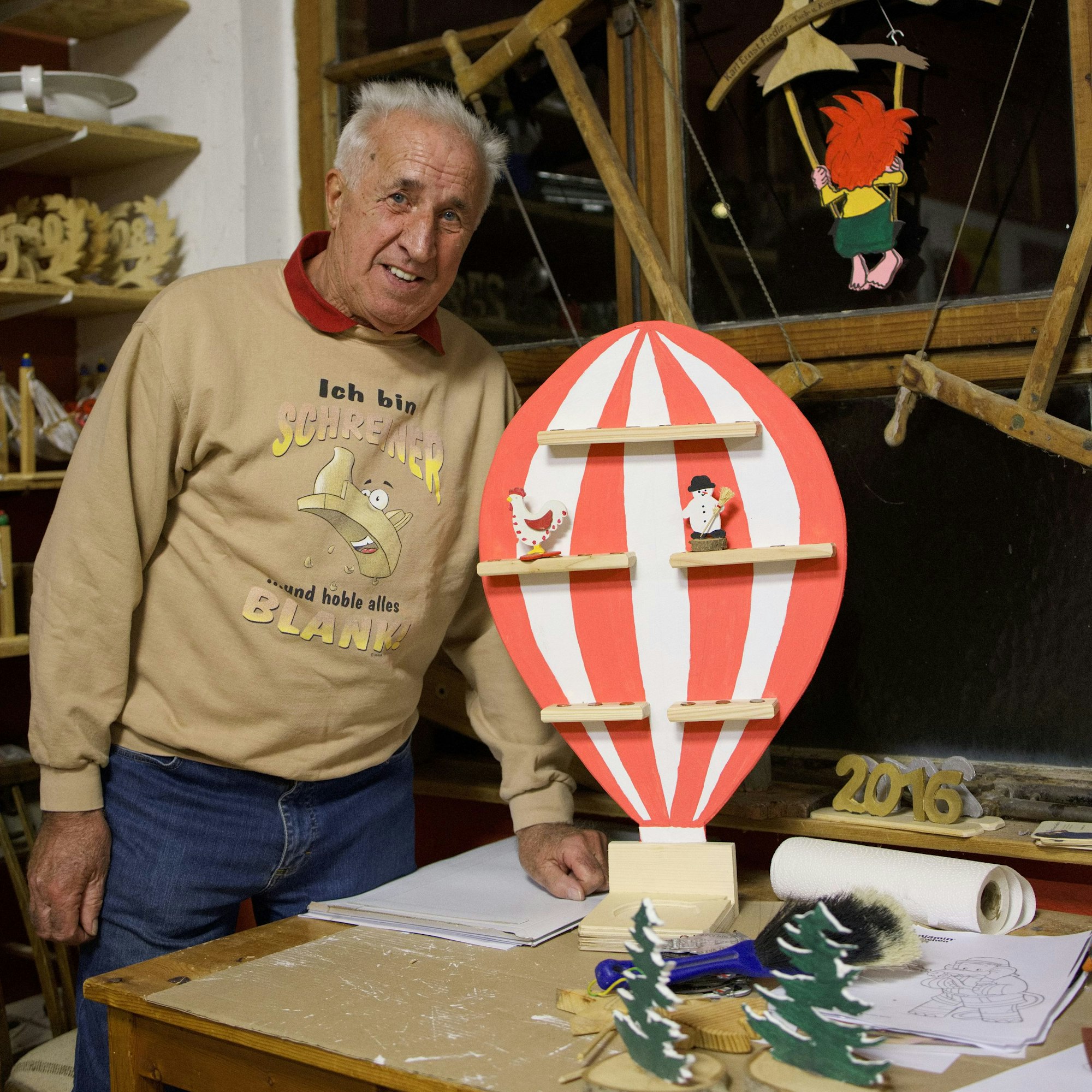 Der Schreiner steht in seiner Werkstatt. Auf dem Tisch steht ein aus Holz geschnittener Heißluftballon, im Hintergrund schaukelt eine Pumuckl-Figur.