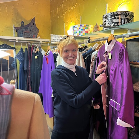 Eine ältere Frau mit kurzem blonden Haar steht in einem Bekleidungsgeschäft und knöpft eine lilafarbene Jacke zu.