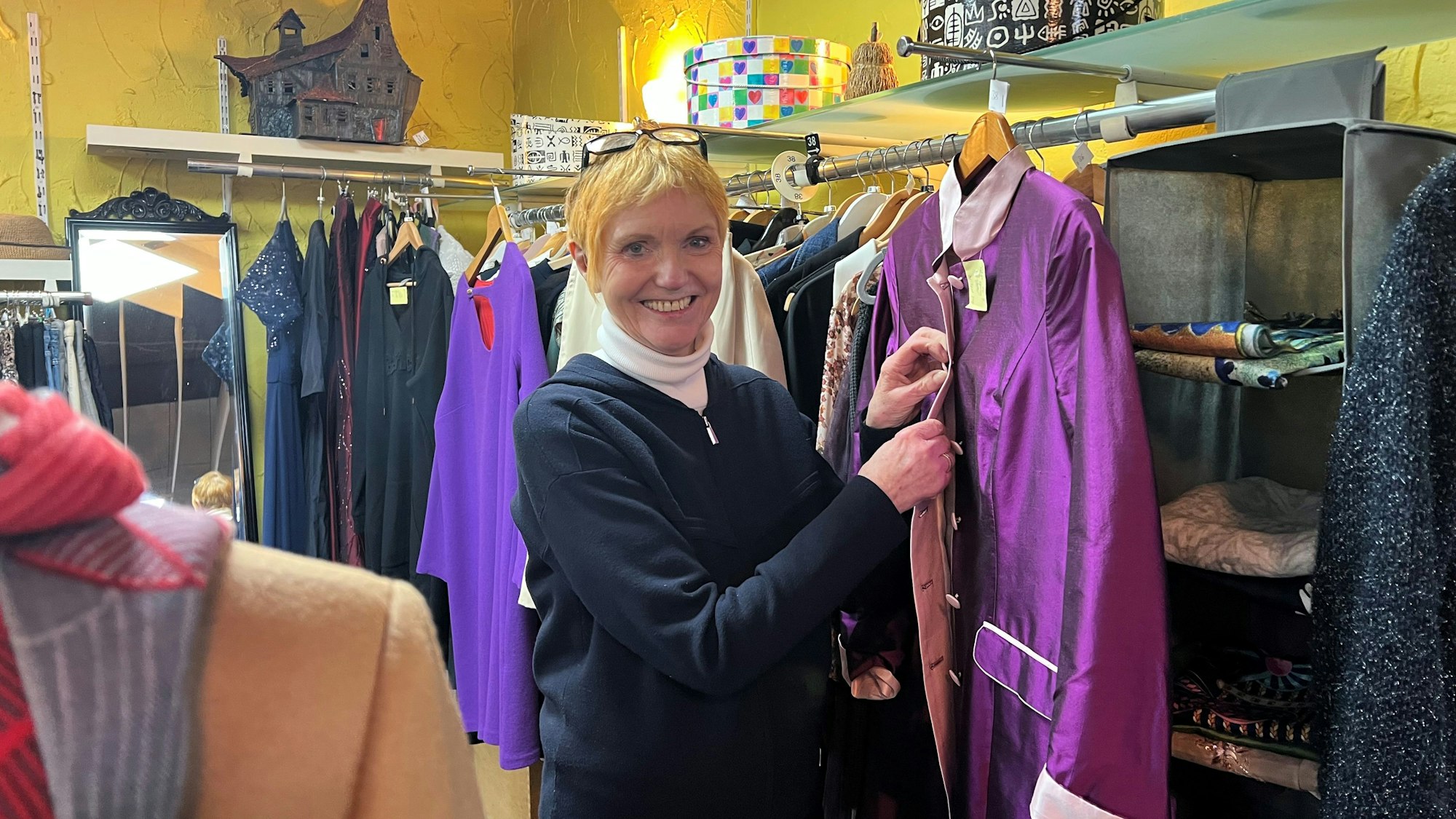Eine ältere Frau mit kurzem blonden Haar steht in einem Bekleidungsgeschäft und knöpft eine lilafarbene Jacke zu.