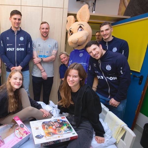 Zwei Mädchen sitzen auf dem Bett im Krankenhauszimmer, während die fünf Handball-Profis und das Maskottchen daneben stehen.
