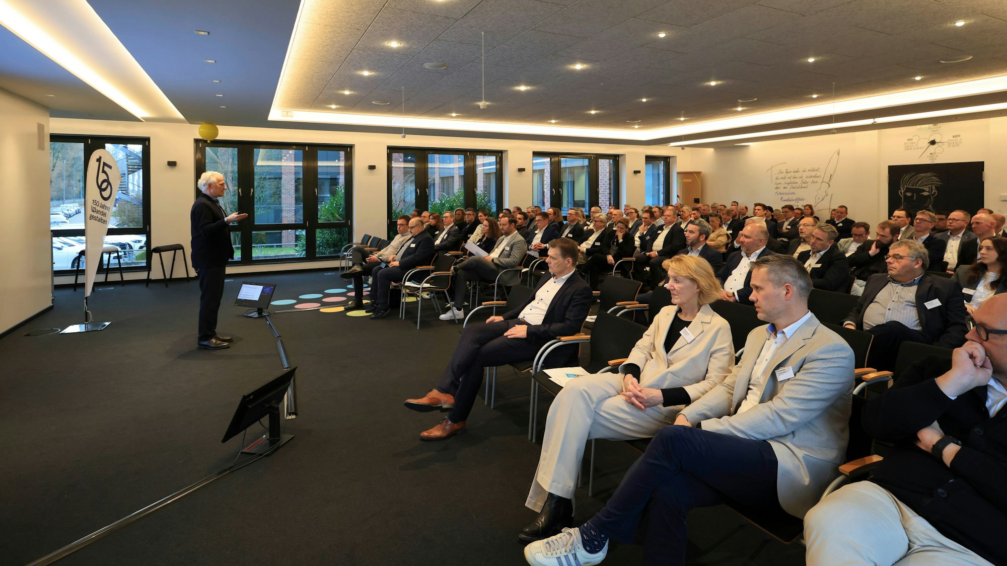 Eine Menschenmenge sitzt geordnet in Sitzreihen und hört einem Redner zu.