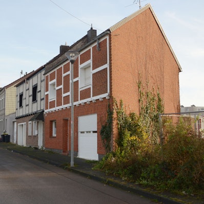 Das Bild zeigt eine Häuserreihe, im Hintergrund geparkte Autos und ein Fúßgänger.