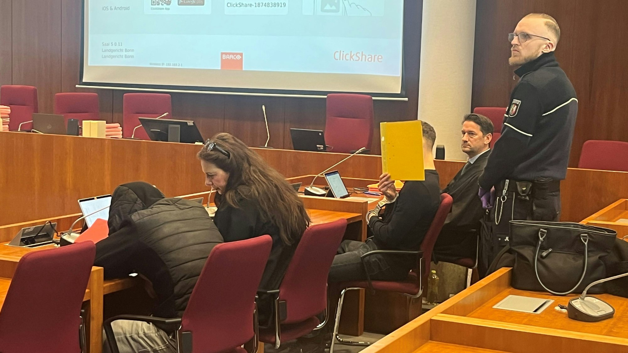 Angeklagte und Anwälte sowie ein Justizbediensteter in einem Gerichtssaal.