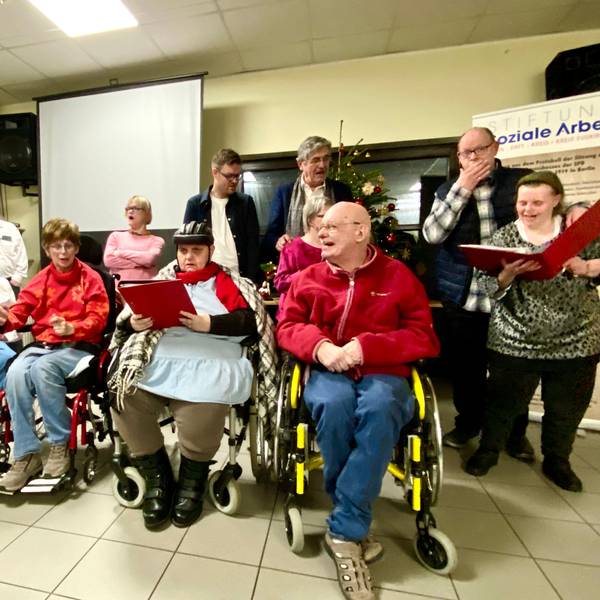 Eine Gruppe von Menschen mit und ohne Behinderung stehen und sitzen beieinander und singen.