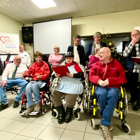 Eine Gruppe von Menschen mit und ohne Behinderung stehen und sitzen beieinander und singen.