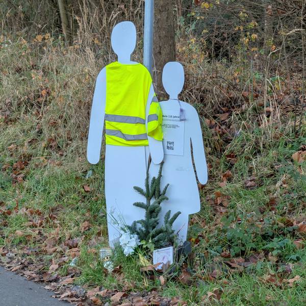 Das Foto zeigt zwei Holzfiguren am Straßenrand, eine davon trägt eine Sicherheitsweste.