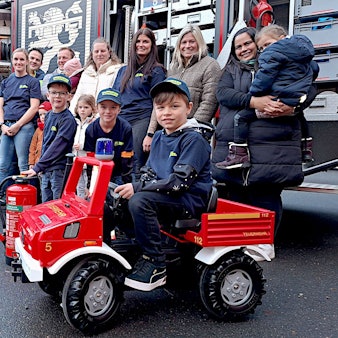 Ein Junge sitzt auf einem Mini-Feuerwehrfahrzeug. Neben ihm stehen weitere Kinder, hinter ihnen einige Erwachsene.