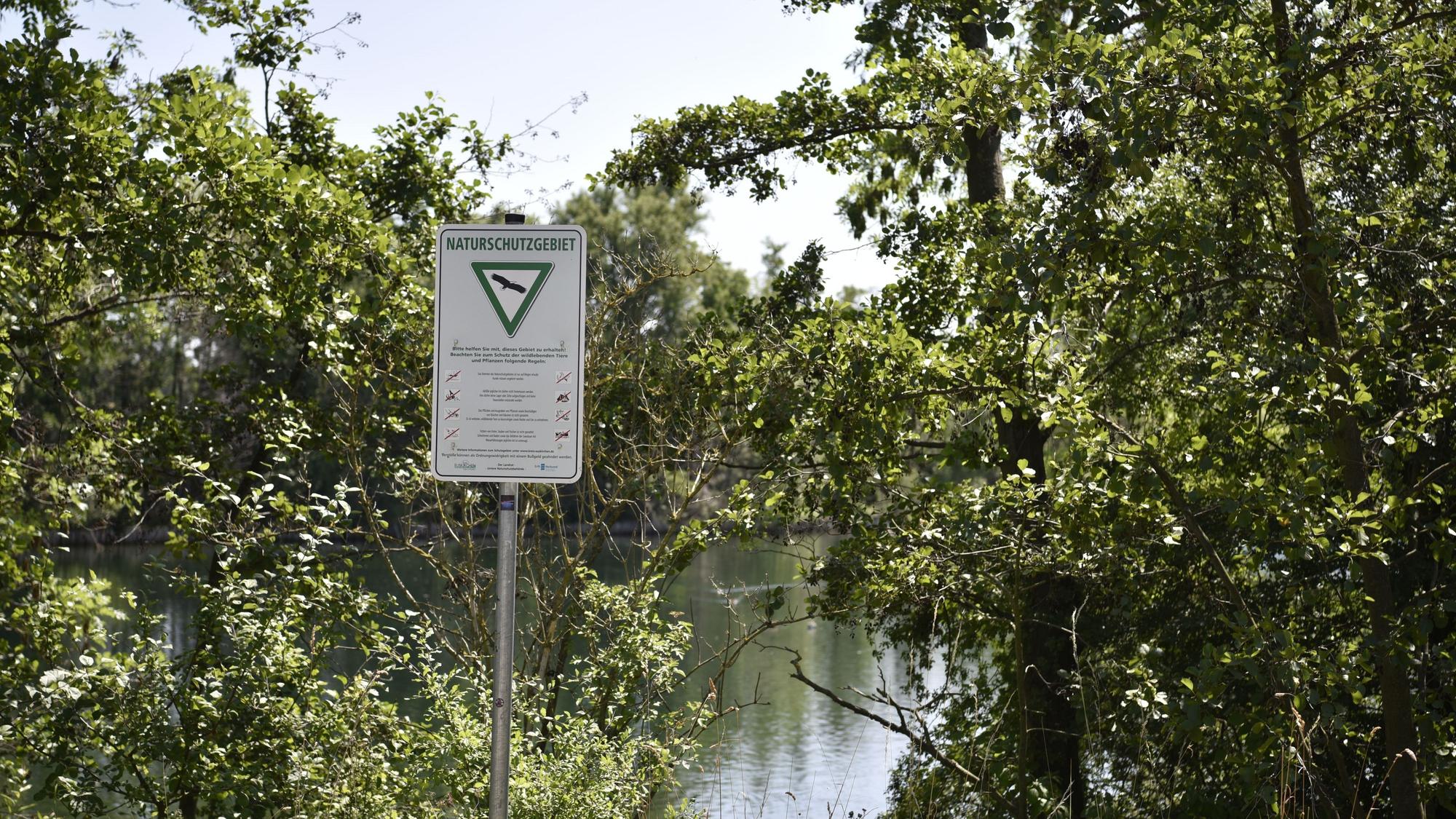 Zwischen Bäumen sieht man Wasser schimmern, davor steht ein Schild, das auf ein Naturschutzgebiet hinweist.