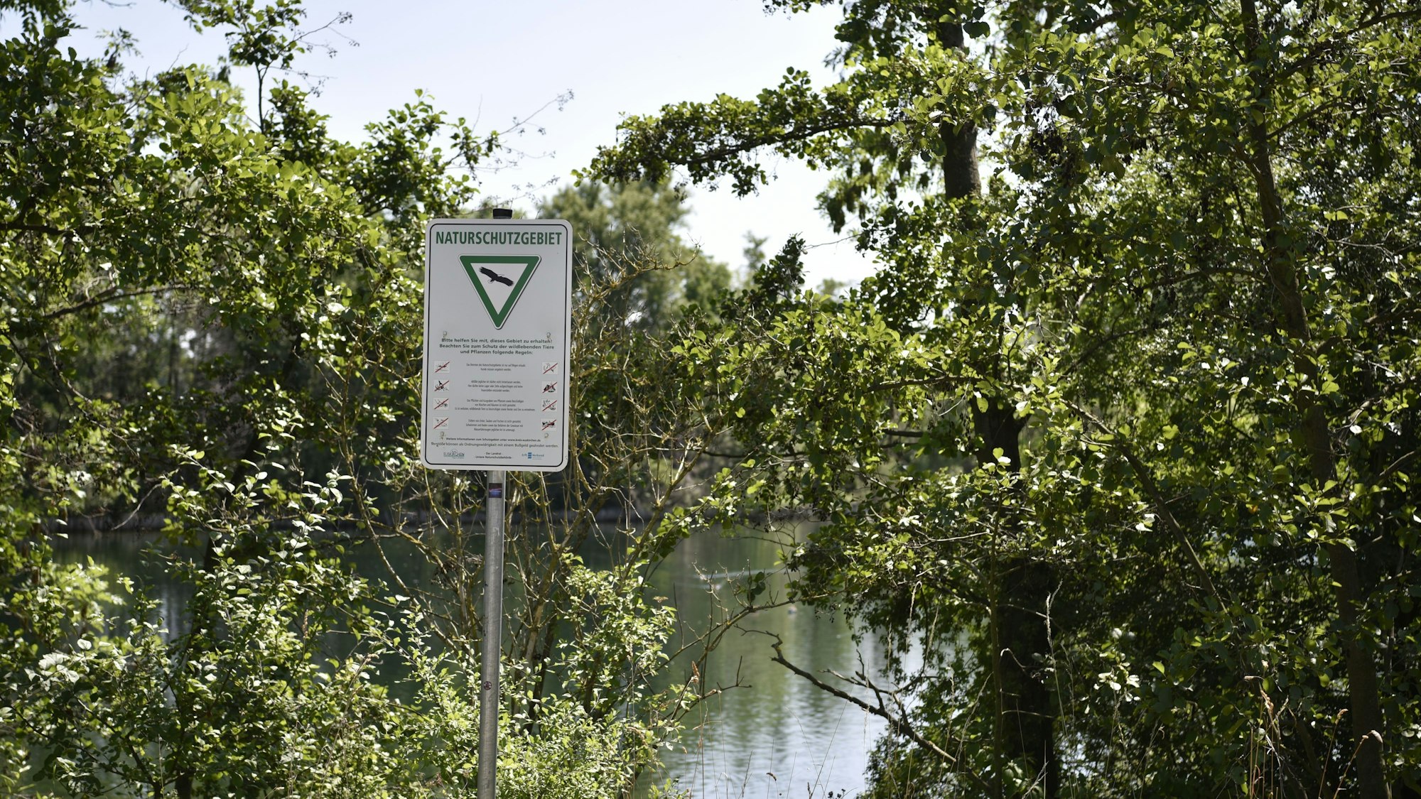 Zwischen Bäumen sieht man Wasser schimmern, davor steht ein Schild, das auf ein Naturschutzgebiet hinweist.