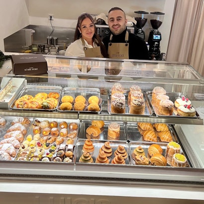 Eine Frau mit langem schwarzem Haar und ein Mann mit kurzem schwarzem Haar stehen hinter einer Gebäcktheke, in der unterschiedliche süße Teilchen liegen.