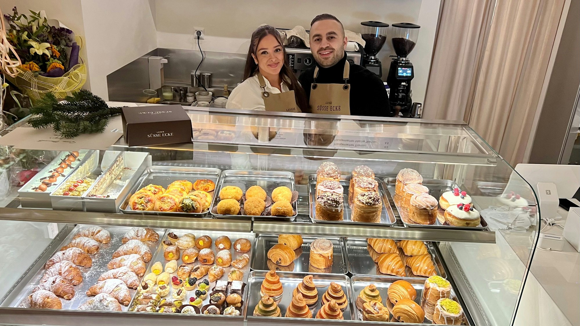 Eine Frau mit langem schwarzem Haar und ein Mann mit kurzem schwarzem Haar stehen hinter einer Gebäcktheke, in der unterschiedliche süße Teilchen liegen.