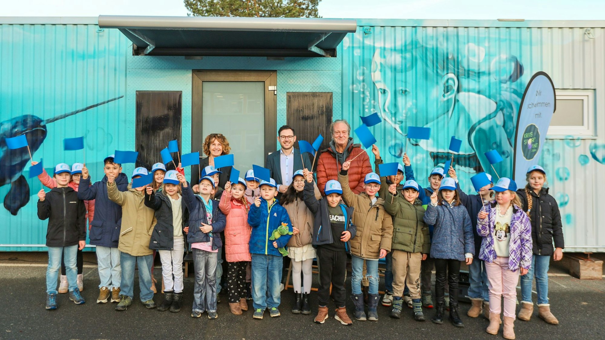 Viele Kinder winken mit blauen Fähnchen, dahinter stehen drei Erwachsene.