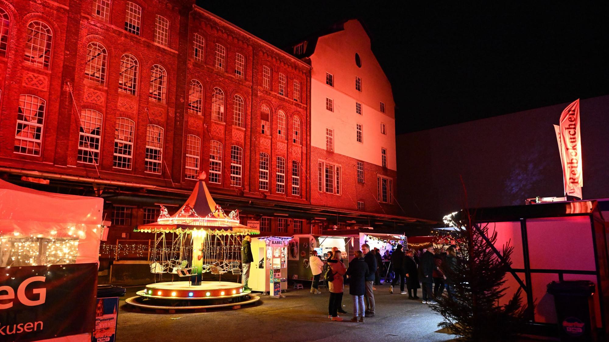 Eine große, rot beleuchtete Hauswand lenkt den Blick auf sich. Davor befindet sich ein kleiner Weihnachtsmarkt.