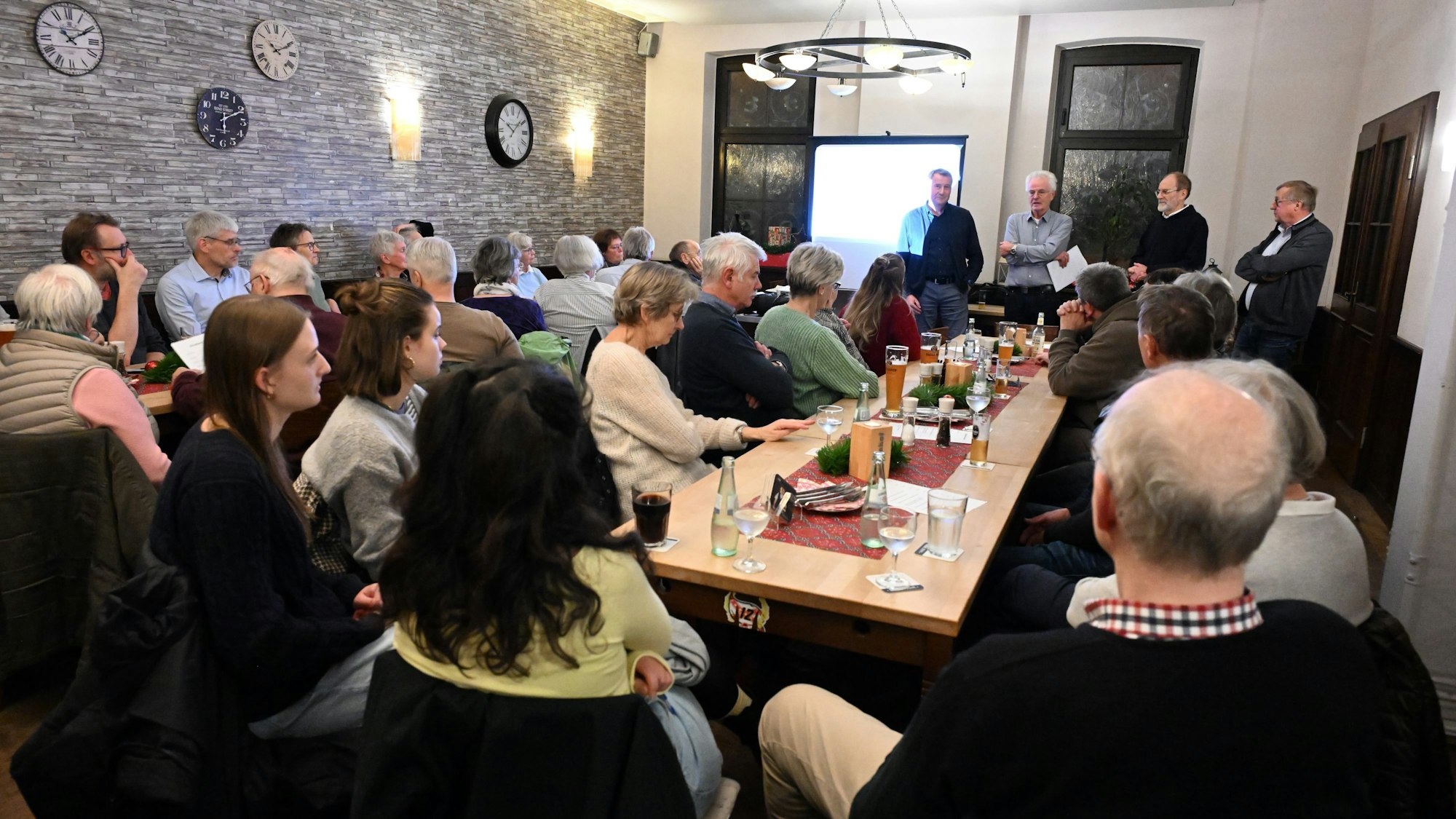 Ein Saal, darin sitzen Männer und Frauen an langen Tischen: vorne stehen vier Männer.
