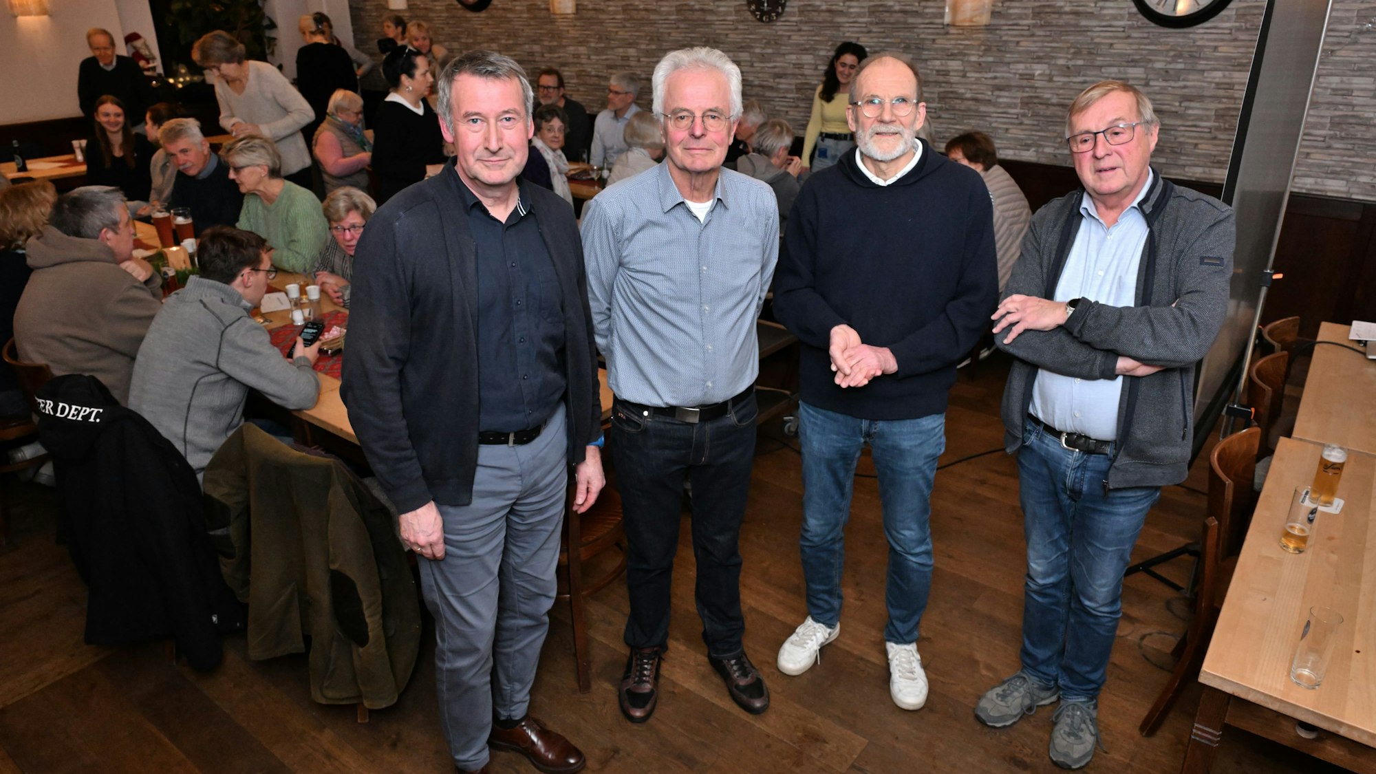 Vier Männer stehen in einem Saal. An langen Tischen sitzen Männer und Frauen.