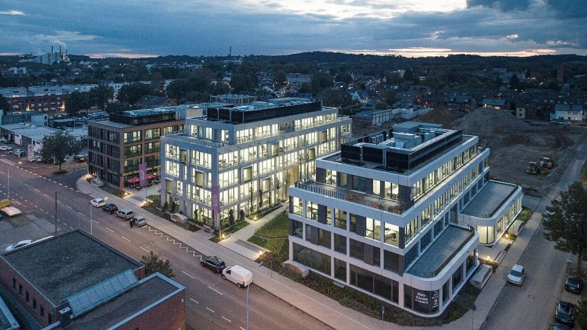 Auf dem Bild ist eine Luftaufnahme von drei erleuchteten Bürogebäuden zu sehen.