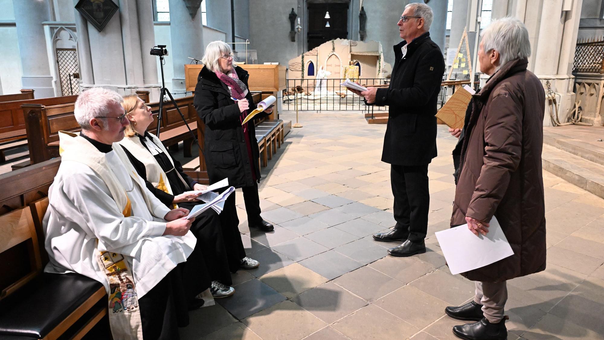 Ein Pfarrer und eine Pfarrerin in Talaren, zwei weitere Frauen und ein Mann stehen in einer Kirche mit Zetteln in der Hand.