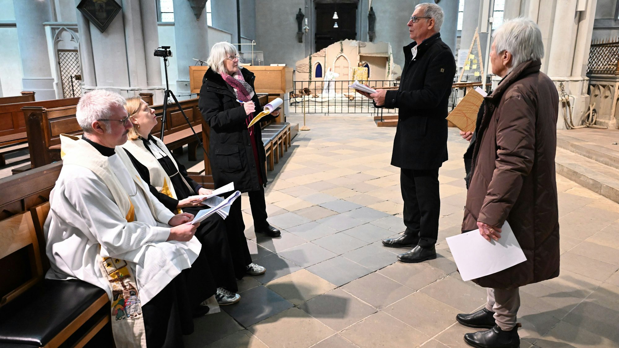 Ein Pfarrer und eine Pfarrerin in Talaren, zwei weitere Frauen und ein Mann stehen in einer Kirche mit Zetteln in der Hand.