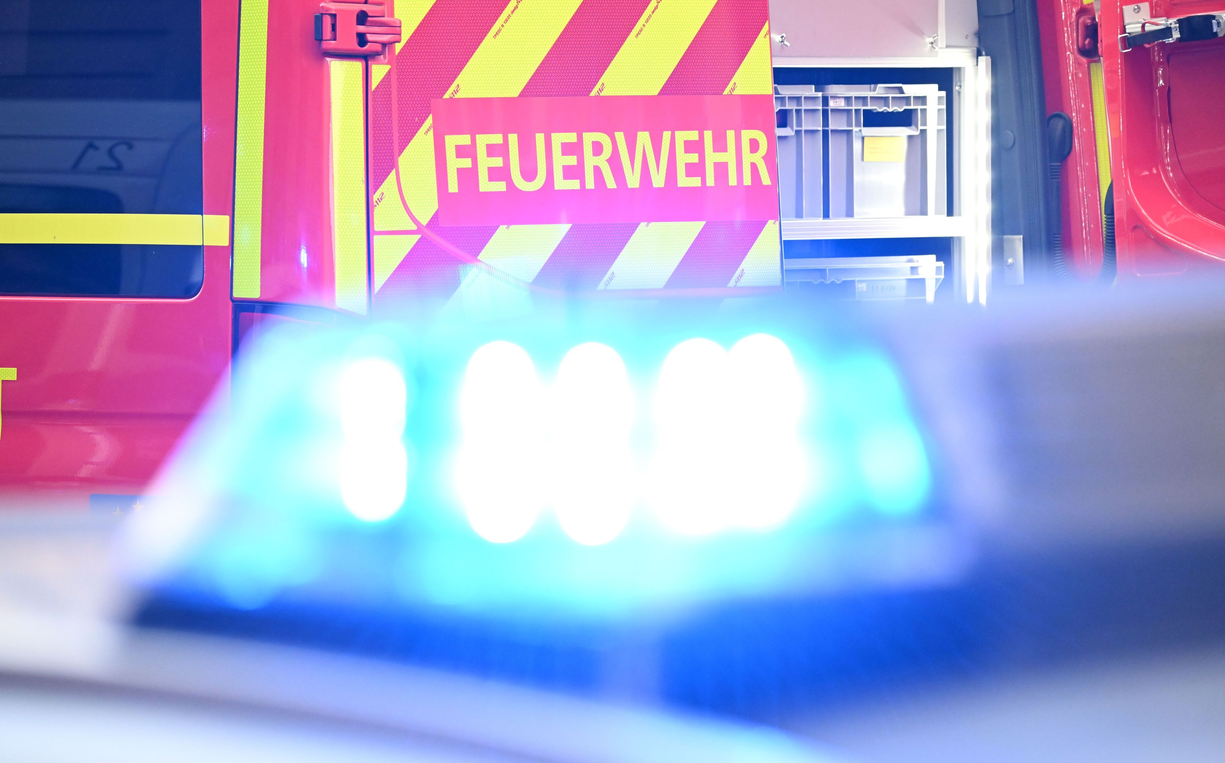 Ein Fahrzeug der Feuerwehr steht bei einem Einsatz auf einer Straße, während im Vordergrund das Blaulicht eines Polizeifahrzeugs zu sehen ist.