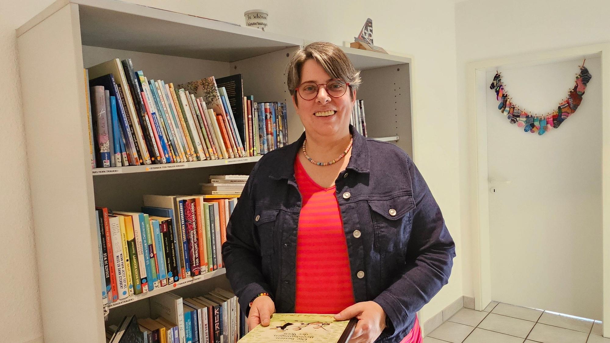 Eine Frau steht in einem Raum und hält ein Buch in der Hand, hinter ihr ist ein Bücherregal.