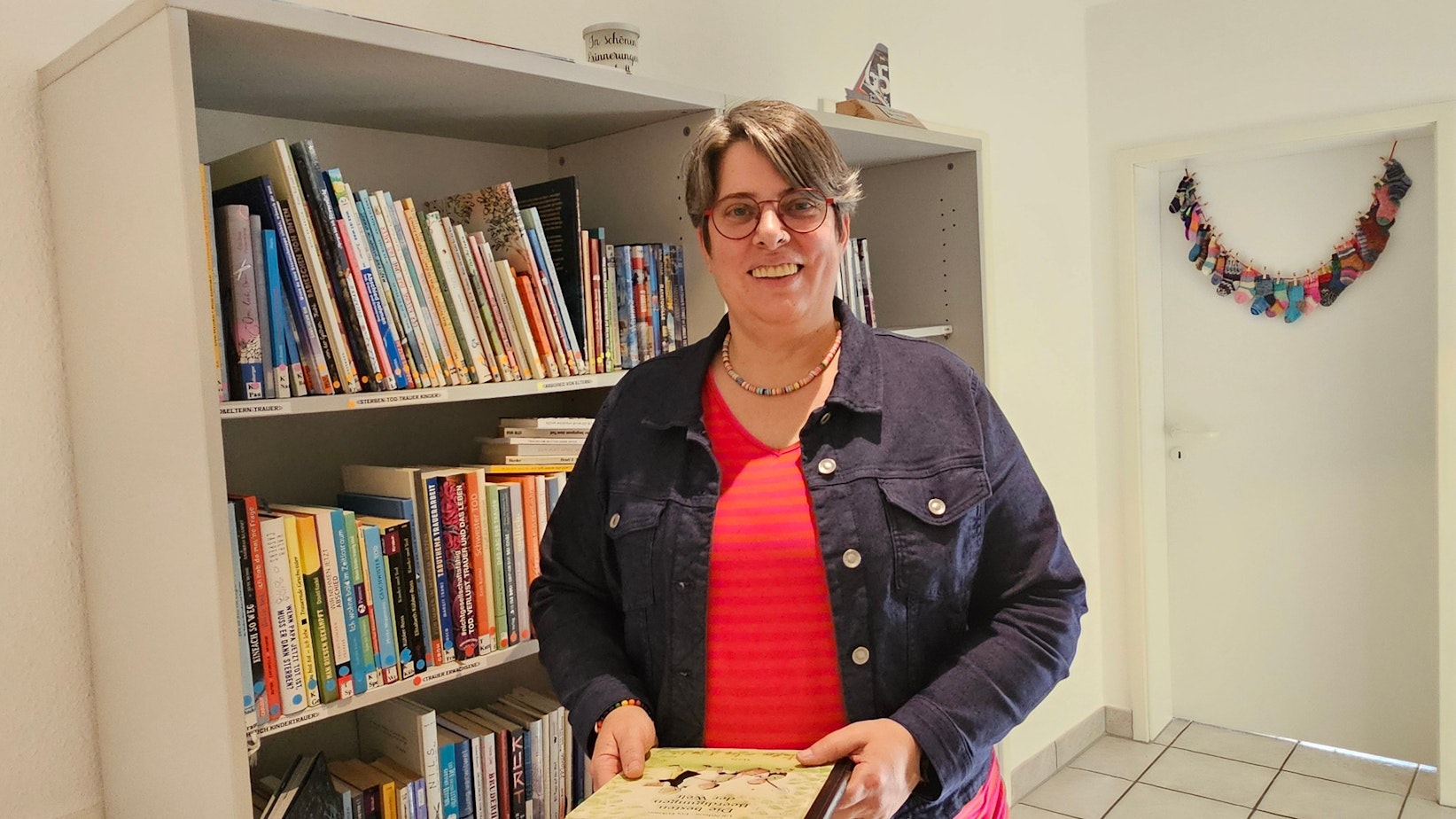 Eine Frau steht in einem Raum und hält ein Buch in der Hand, hinter ihr ist ein Bücherregal.