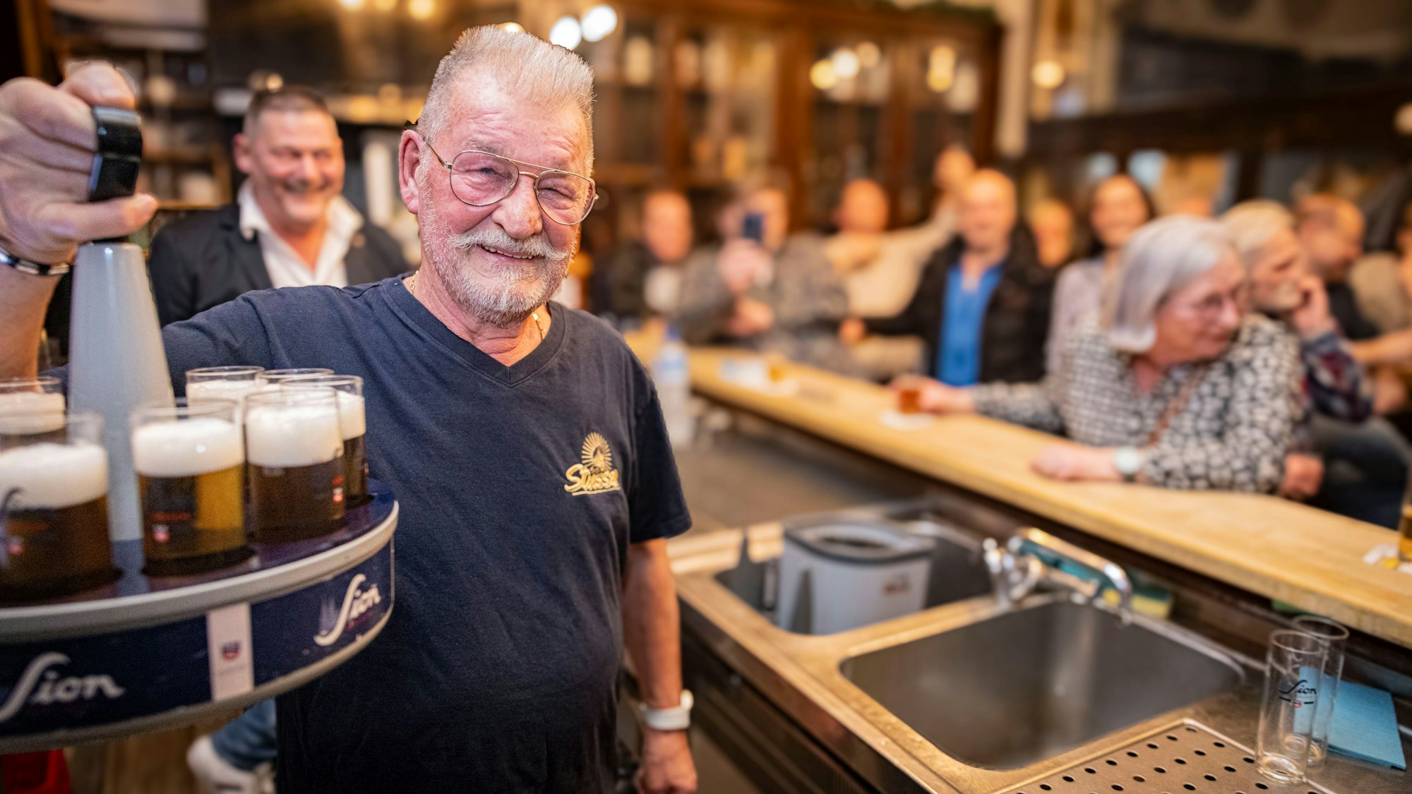 Köbes Karl Heinz Fischer feiert seinen Abschied im Brauhaus Stüsser. Er lächelt in die Kamera und hat einen Kölschkranz in der Hand.