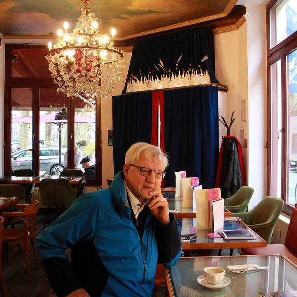 Ein Mann in blauem Anorak sitzt an einem Hochtisch in einem Café. An der Decke hängt ein großer Kristall-Kerzenleuchter.