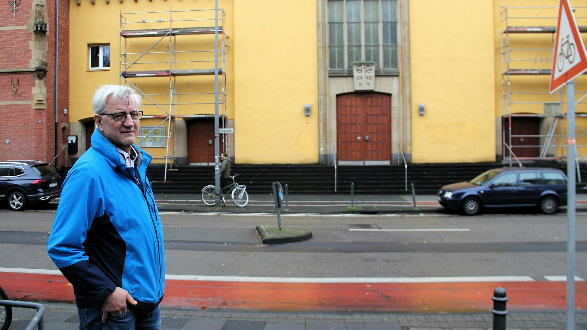Ein Mann im blauen Anorak steht an einer Straße, auf der andren Seite ist ein gelbgetünchter Kirchenbau zu sehen.