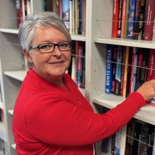 Auf dem Bild ist eine Frau in einem roten Oberteil vor einem Bücherregal zu sehen.