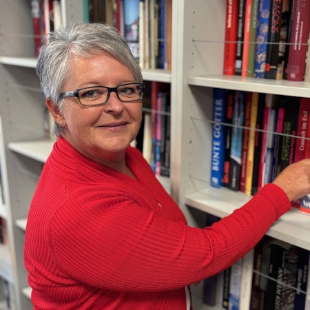 Auf dem Bild ist eine Frau in einem roten Oberteil vor einem Bücherregal zu sehen.