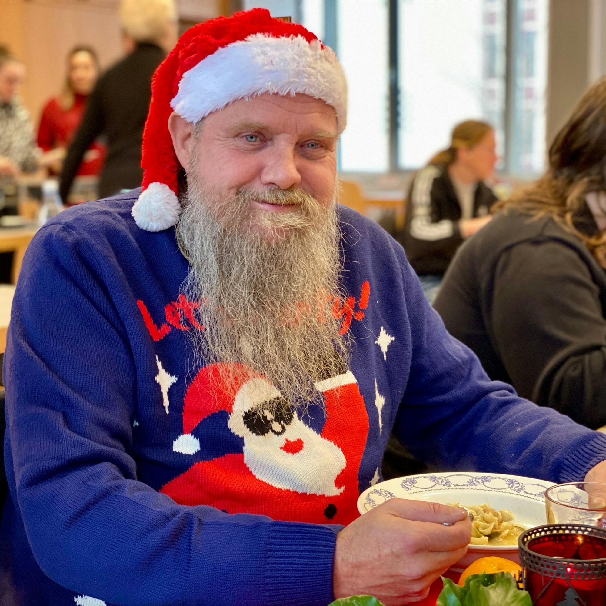 Ein Gast der Feier sitzt beim Essen. Auf dem Kopf trägt er eine Weihnachtsmütze, dazu einen blauen Weihnachtspullover. Der Mann hat einen langen grauen Bart und lächelt freundlich in die Kamera.