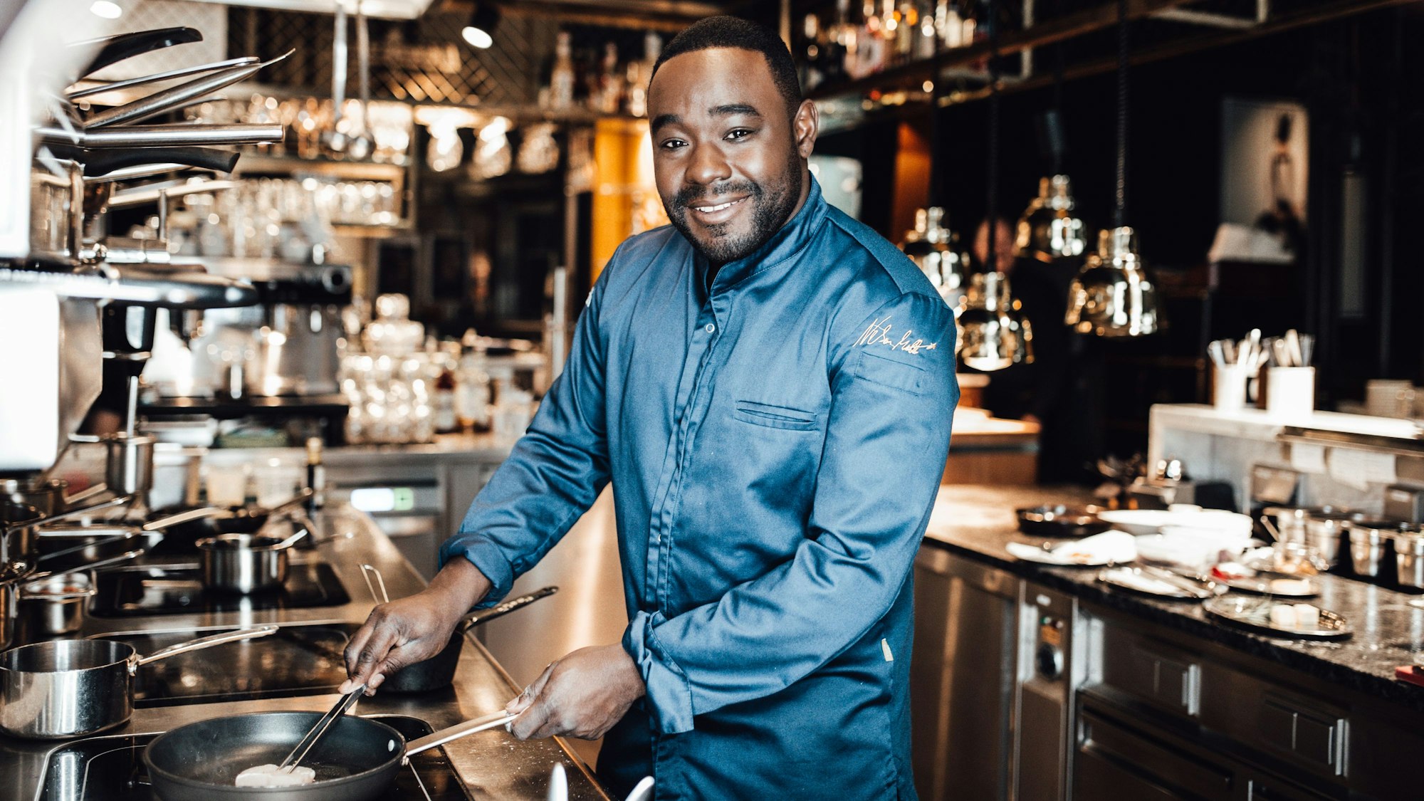 Ein junger Mann steht am Herd mit einer Pfanne und lächelt.