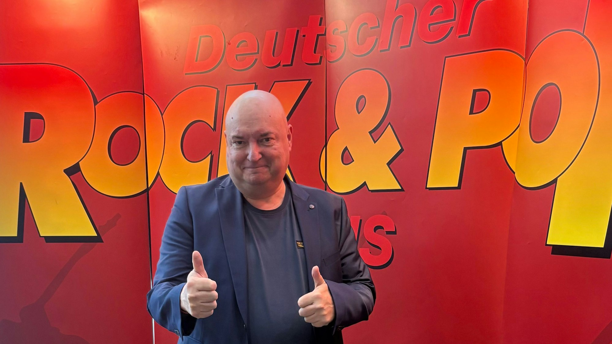 Ein Mann steht vor einem roten Plakat mit der Aufschrift „Deutscher Rock-&-Pop Preis“.