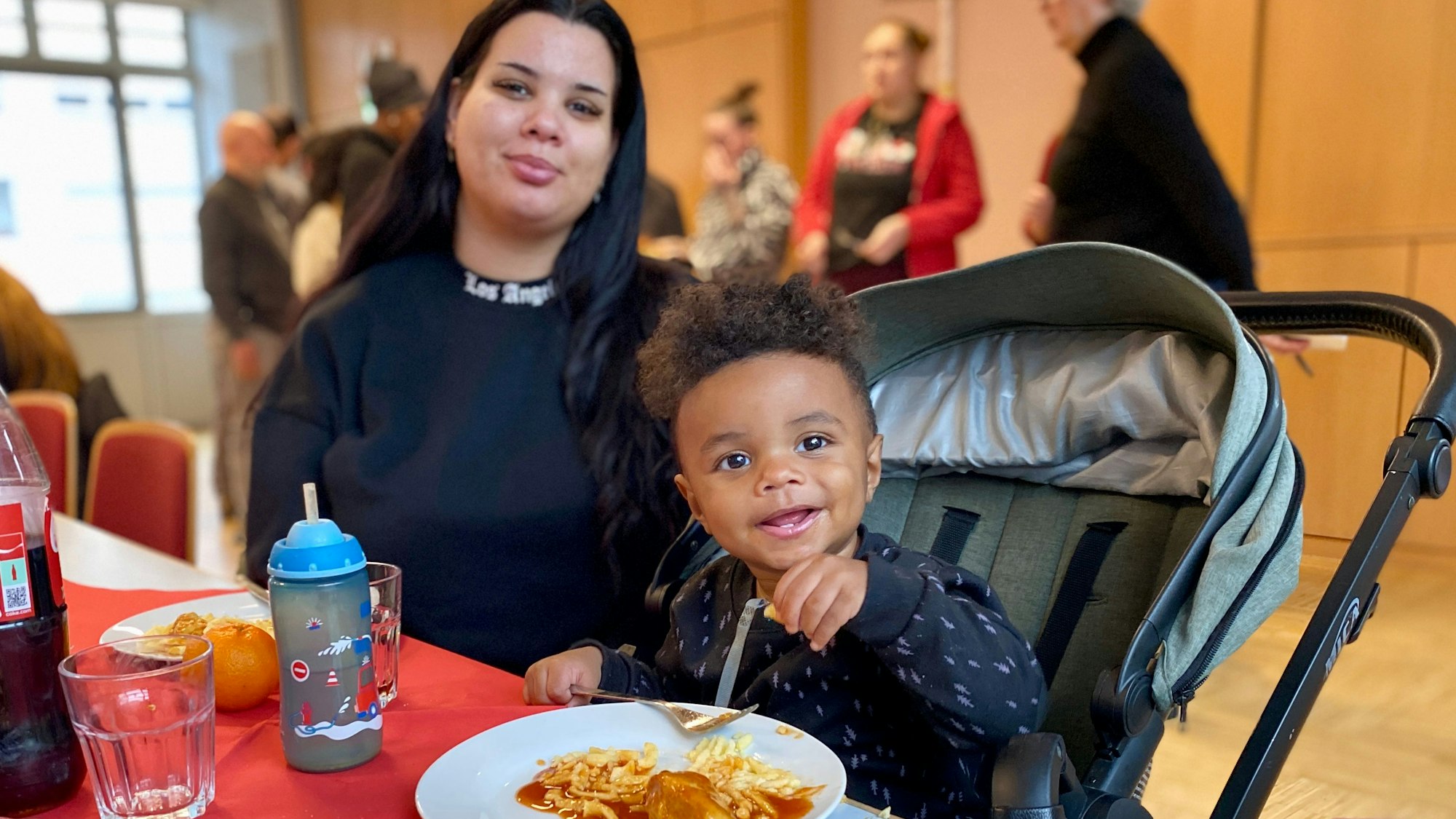 Eine Mutter sitzt am Tisch, daneben ihr kleiner Sohn im Kinderwagen. Beide haben volle Teller vor sich stehen.