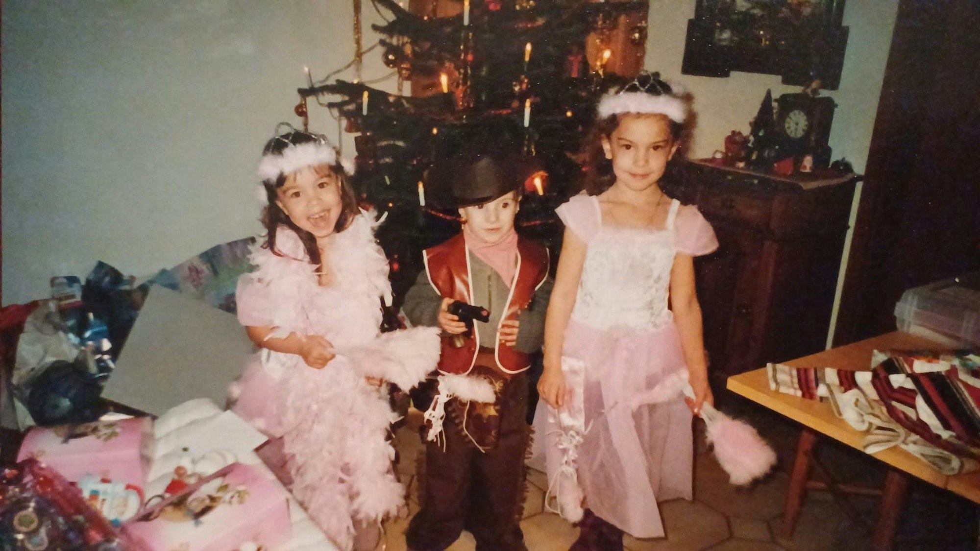 Zwei Mädchen und ein Junge stehen vor einem Weihnachtsbaum. Die Mädchen tragen Prinzessinnenkostüme und der Junge ein Cowboy-Kostüm.