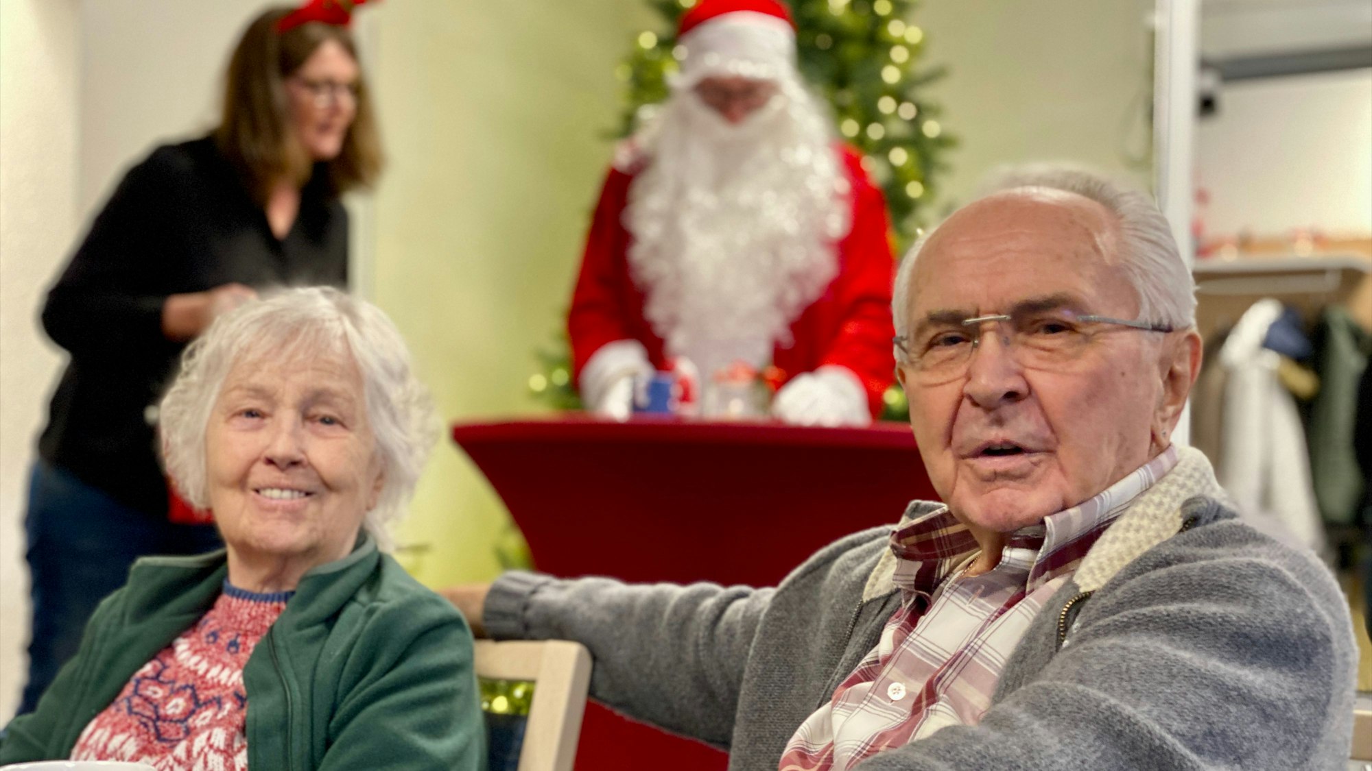 Das Bild zeigt zwei Senioren im Vordergrund. Im Hintergrund ist ein Weihnachtsmann zu sehen.