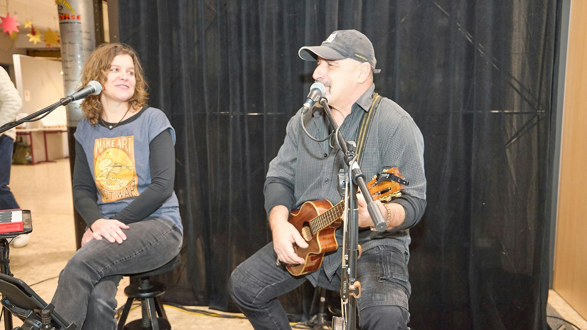 Das Foto zeigt zwei Musiker auf Stühlen sitzend am Mikrofon. Vassil Svechtarov spielt auf einer Gitarre und singt, Inga Beitz-Svechtarov schaut ihn an.