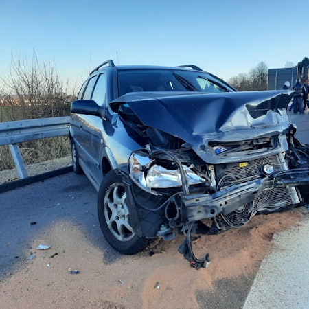 Ein blaues Auto mit demolierter Frontpartie, im Hintergrund ein Einsatzfahrzeug der Feuerwehr.
