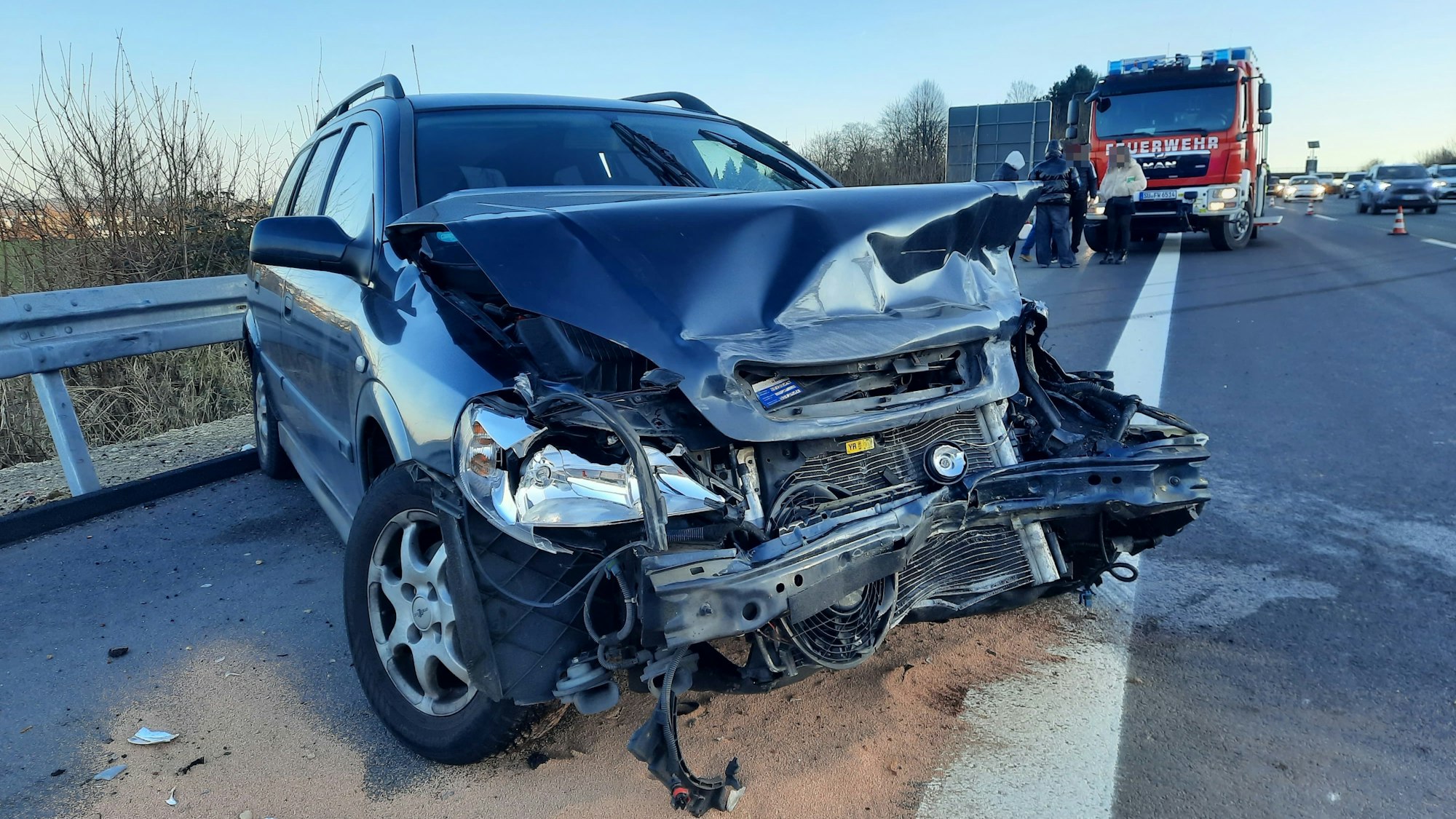 Ein blaues Auto mit demolierter Frontpartie, im Hintergrund ein Einsatzfahrzeug der Feuerwehr.