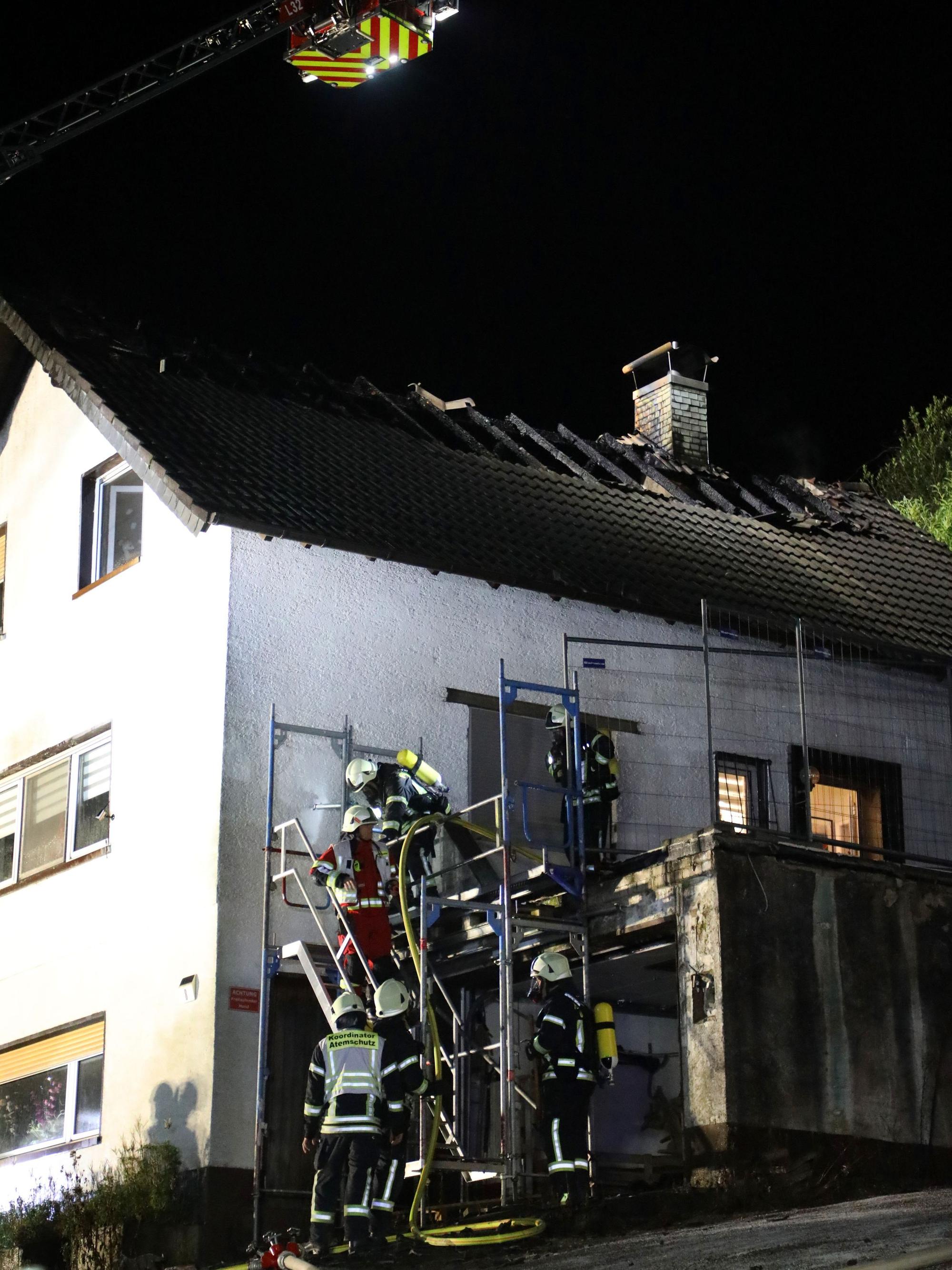 Feuerwehrleute steigen über ein Gerüst zu einem Haus hinauf. Das Dach des Hauses ist abgebrannt.