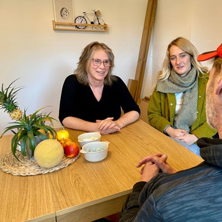 Maria Surges-Brilon und Gina Raths sitzen am Tisch und sind im Gespräch mit dem Bewohner, den man nur von hinten sieht.