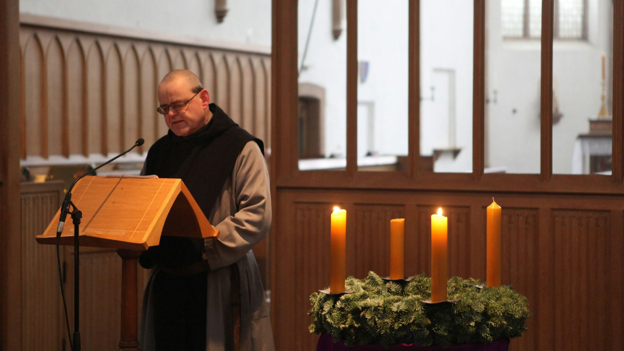 Ein Mönch steht am Lesepult. Im Vordergrund sieht man einen Adventskranz, auf dem zwei Kerzen brennen.
