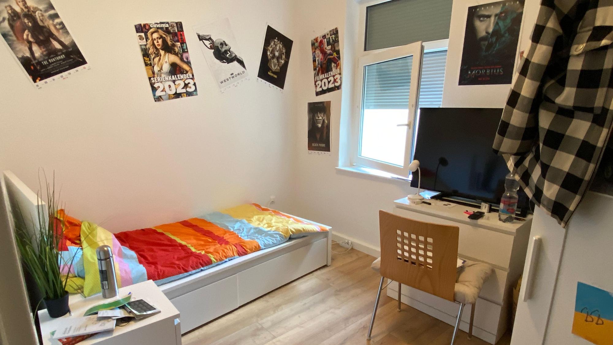 Ein Blick in eines der WG-Zimmer, der Boden ist aus hellem Holz, die Möbel sind weiß, an den Wänden hat der Bewohner Filmplakate gehängt.