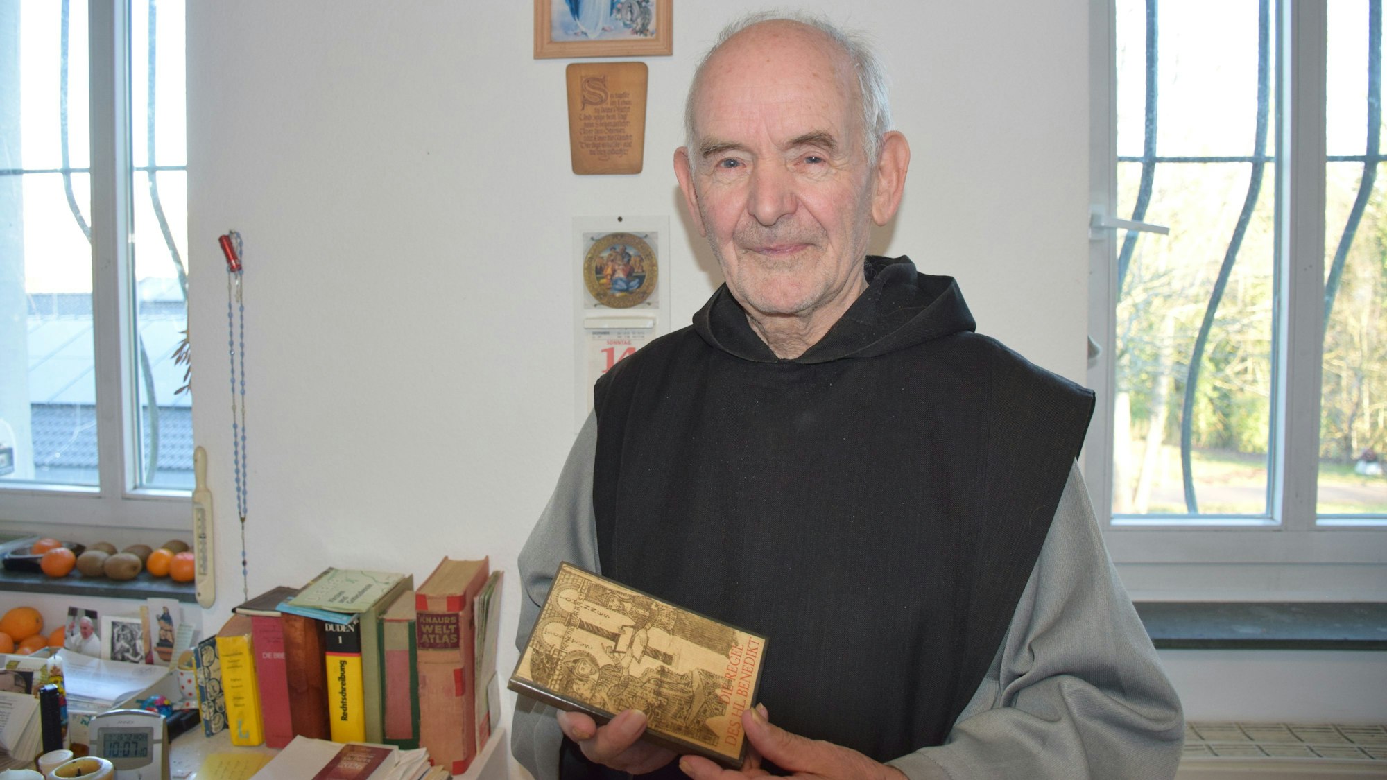 Bruder Clemens Maria, der letzte Mönch aus dem Kloster Mariawald, lächelt freundlich in die Kamera. In den Händen hält er ein kleines Buch.