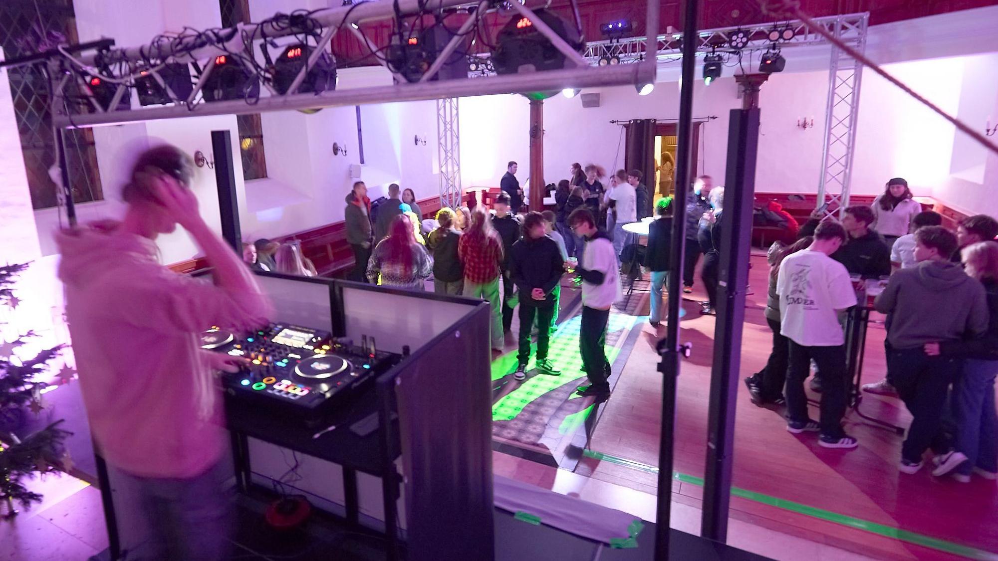Der Kircheninnenraum ist bunt beleuchtet, etliche junge Menschen tanzen oder stehen an Biertischen zusammen. Im Vordergrund sieht man den DJ an seinem Pult.