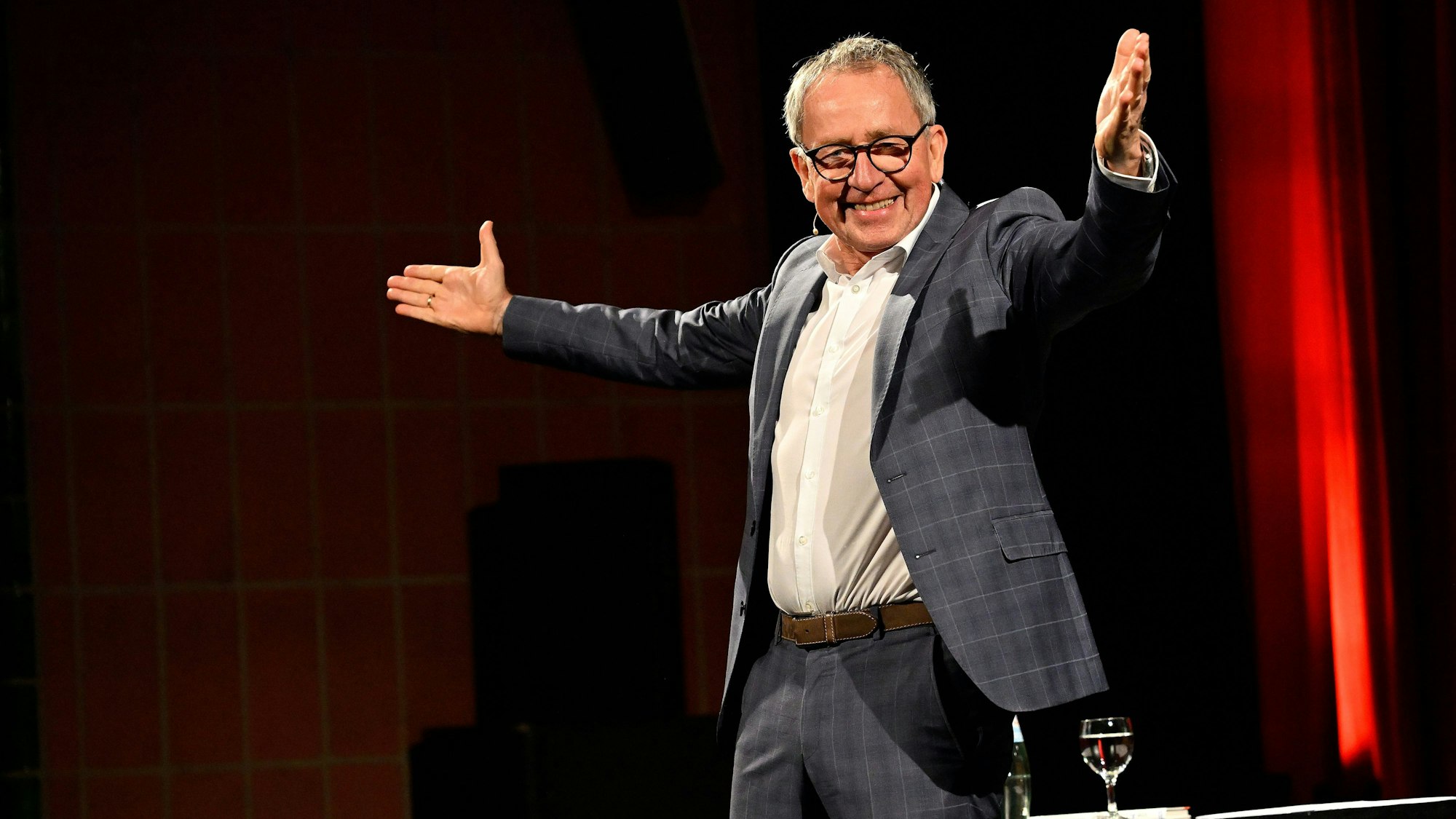 Ein Mann in einem dunklen Karoanzug steht auf einer Bühne mit ausgebreiteten Armen und lächelt.