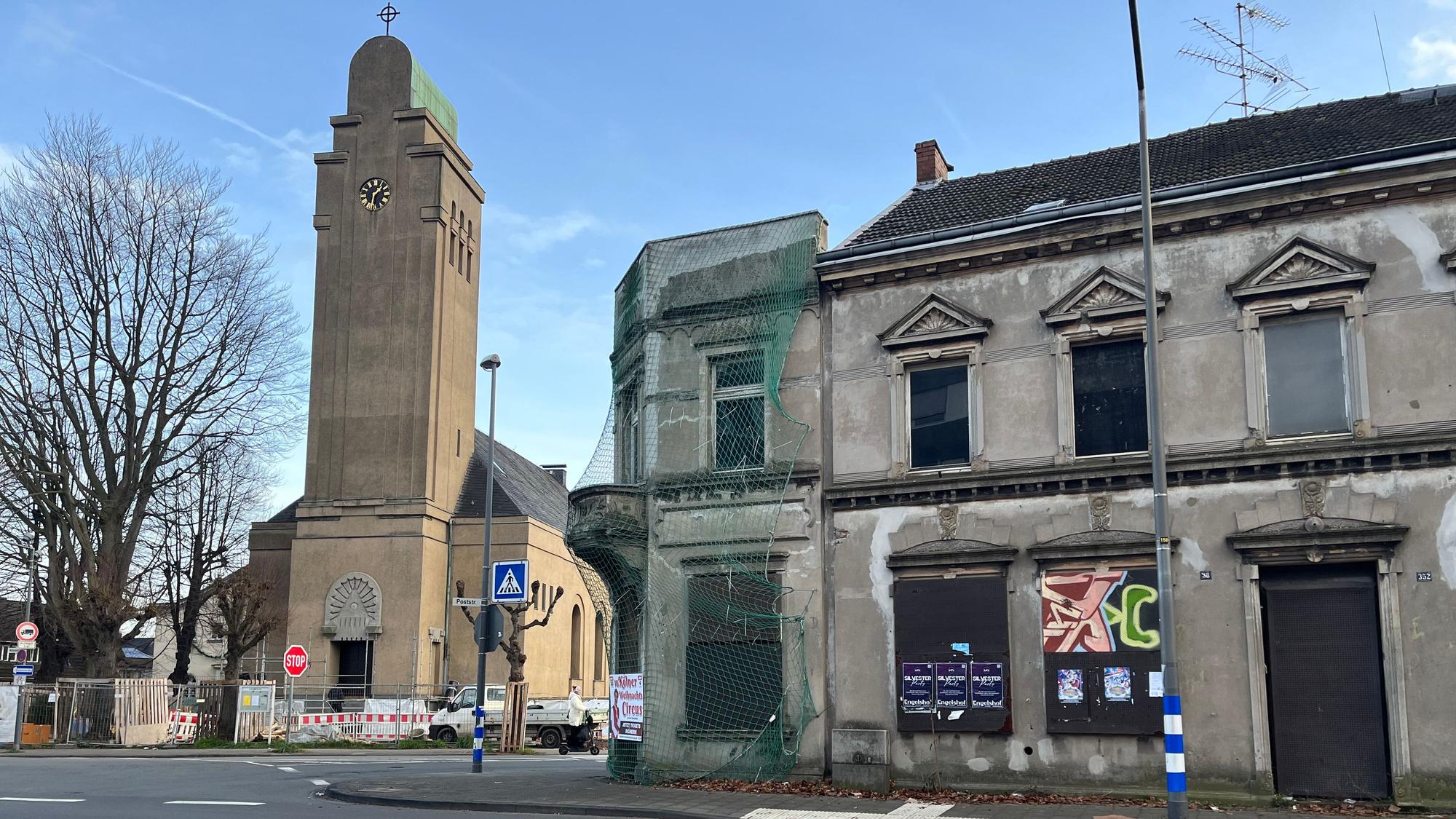 An einer Straße stehen eine Kirche und ein marodes dreistöckiges Haus, das teils von einem Schutznetz umgeben ist.