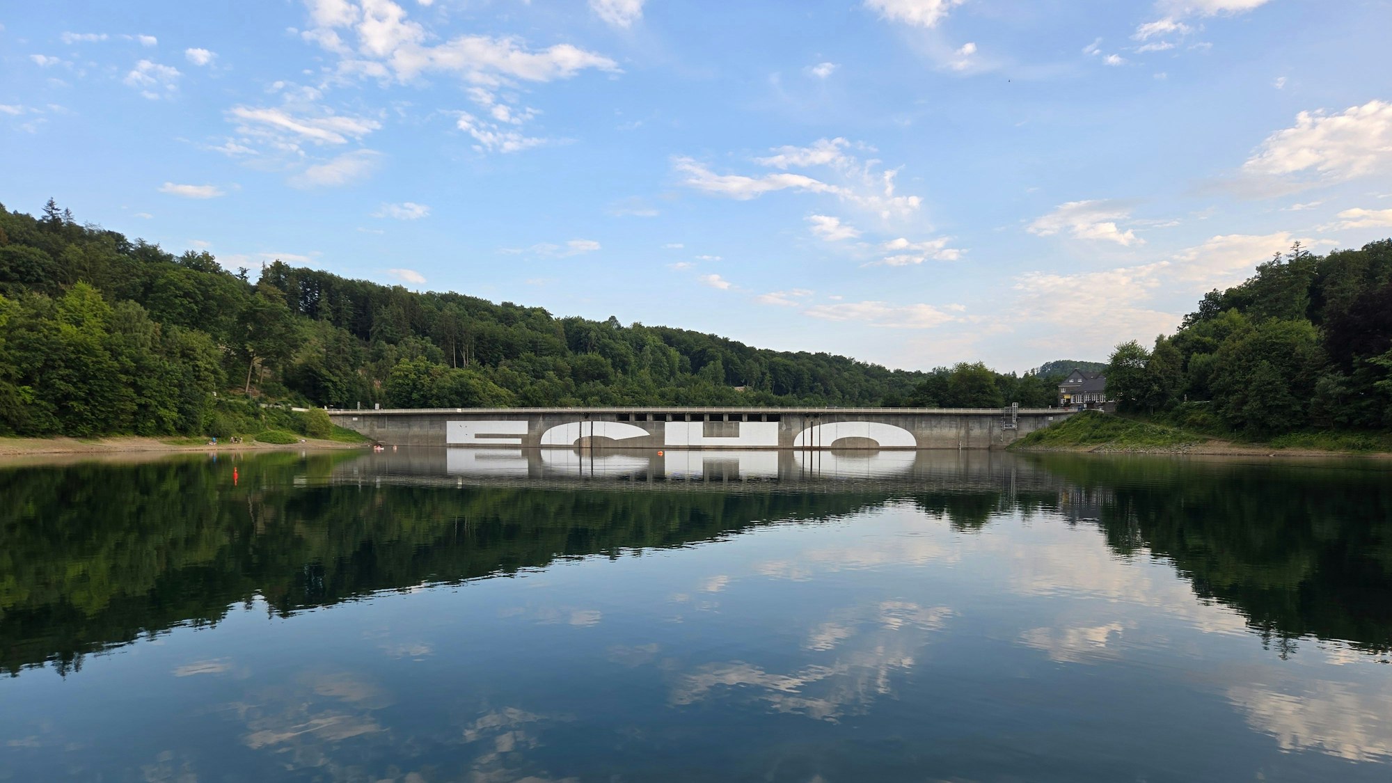 Im Rahmen der Regionale 2025 wird ein großer Schriftzug auf die Mauer der Aggertalsperre gemalt. Das „Echo“ ist erst als Spiegelung vollständig.