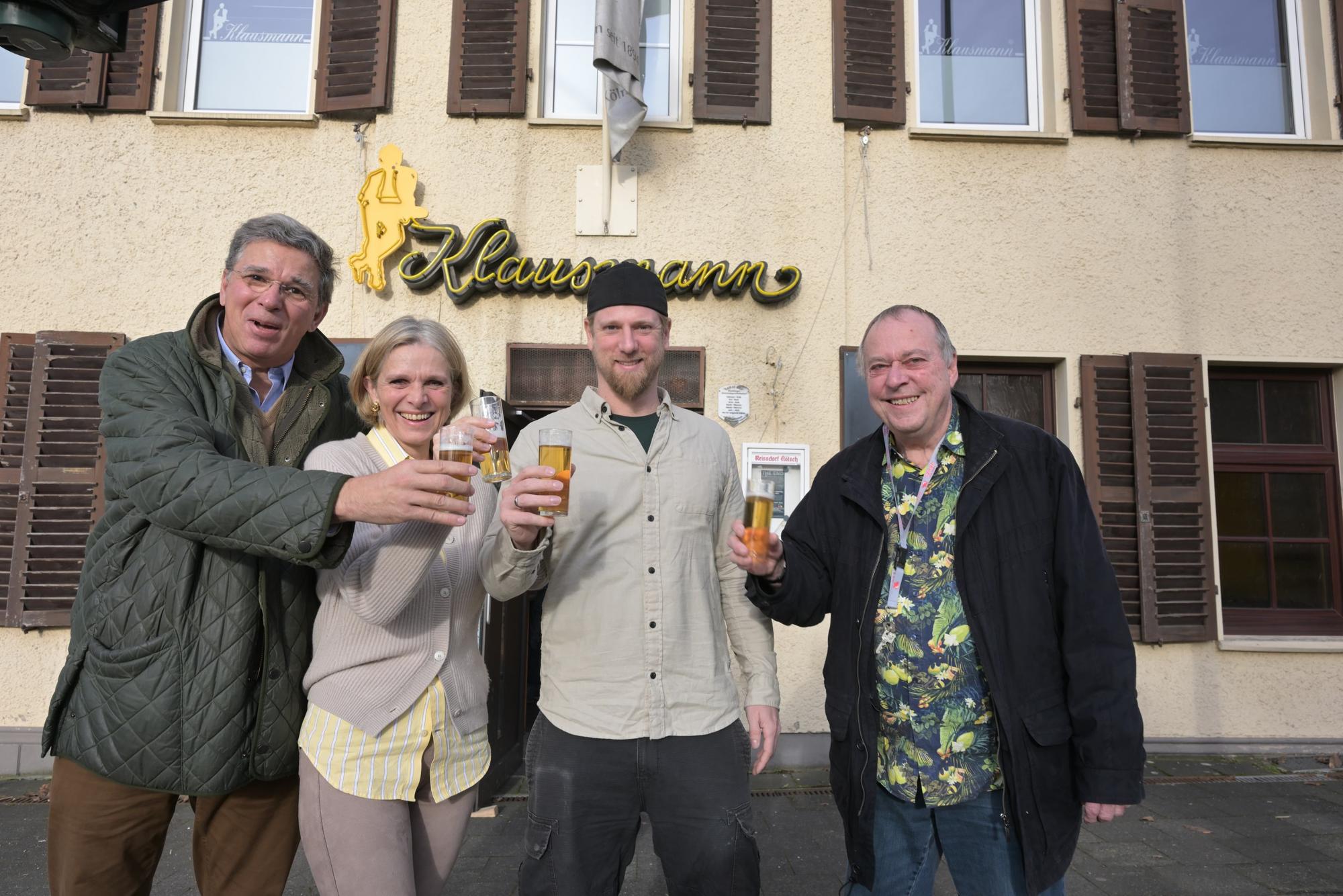 Drei Männer und eine Frau halten ein Glas Kölsch vor sich und lächeln.