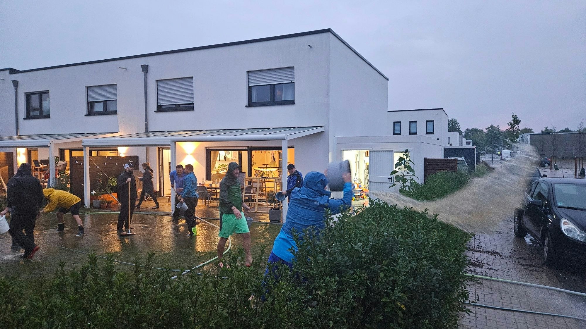 Zu sehen sind Menschen in Regenjacken, die Wasser aus einem Garten schippen.